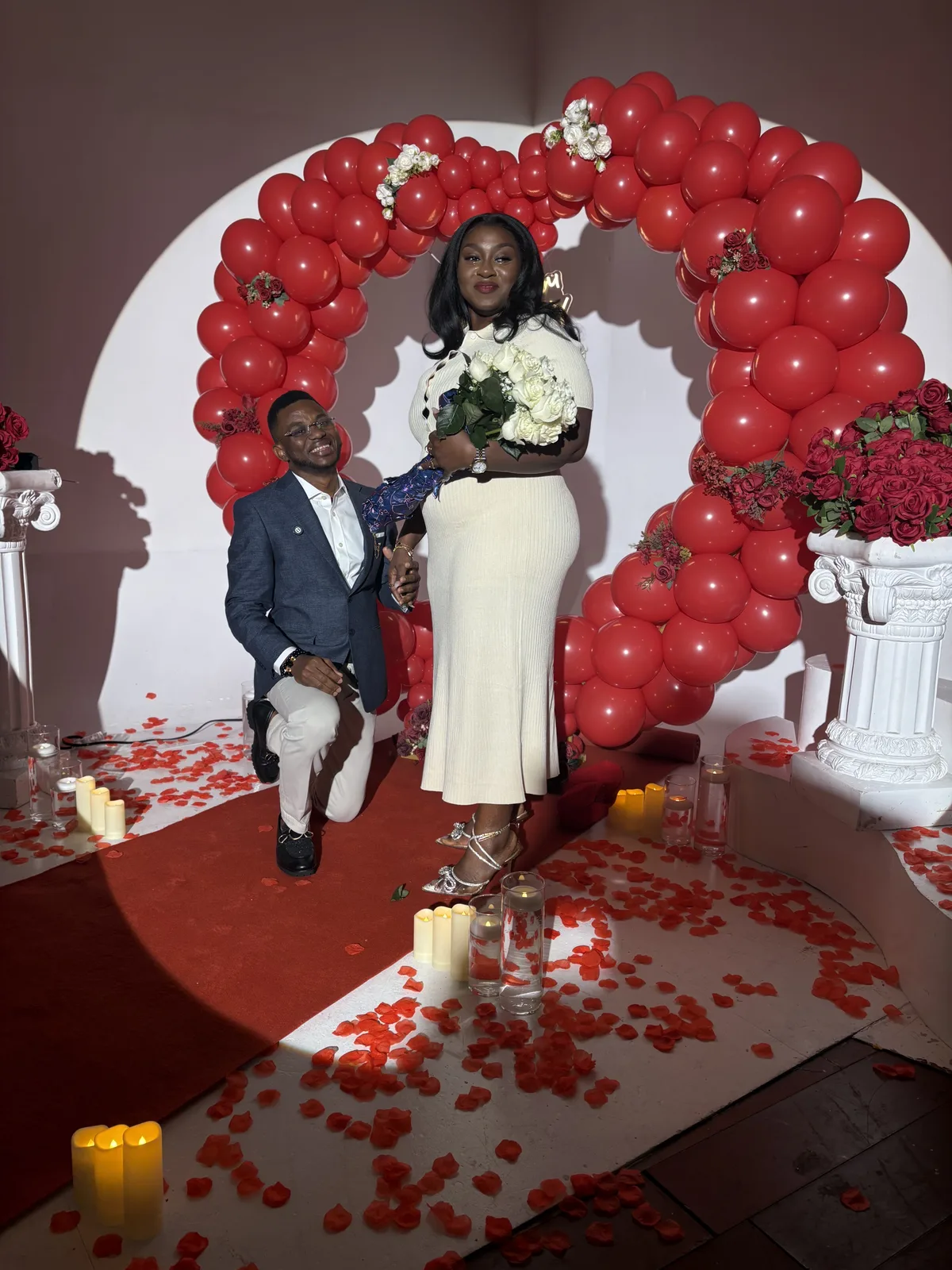 The proposal: a couple beneath a heart-shaped arch of red balloons surrounded by candles and rose petals
